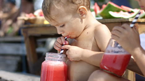 Camera-Focuses-On-A-Wet-Boy-Without-Clothes-On-The-Beach-While-He-Drinks-A-Watermelon-Juice-With-A-Straw-Accompanied-By-His-Sister