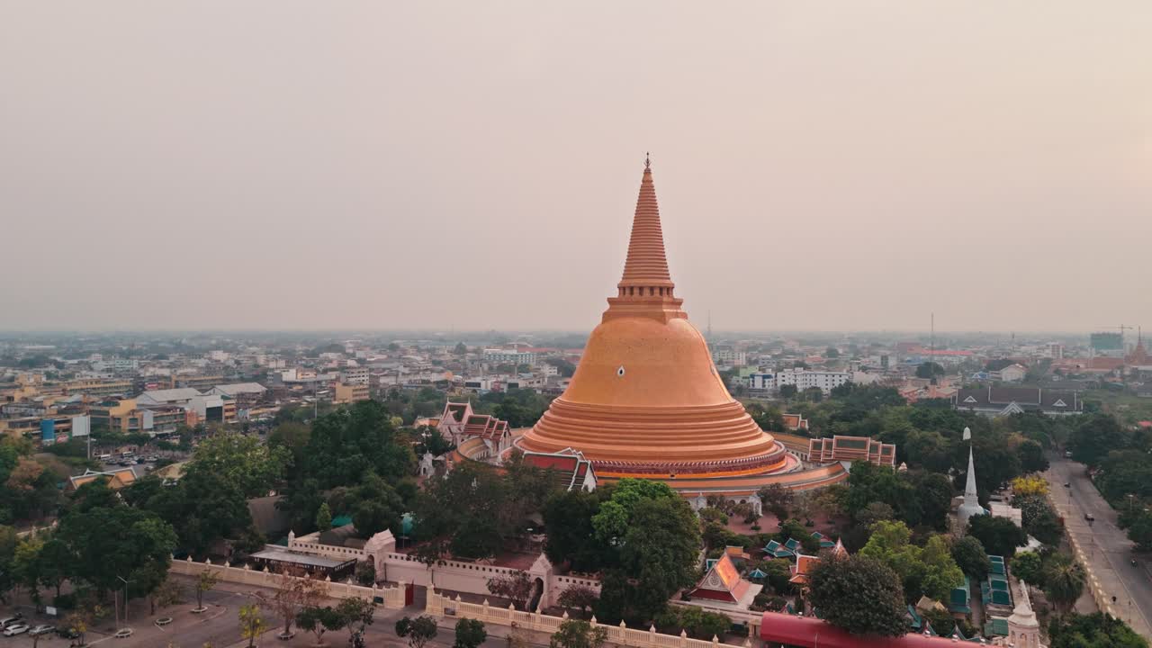 Volo fluido di drone che panoramica a sinistra, catturando un'enorme stupa dorata che si erge sopra il centro urbano, mentre un maestoso monumento buddista domina lo skyline della città in Thailandia sotto un cielo grigio e velato durante un'alba serena e rossa.