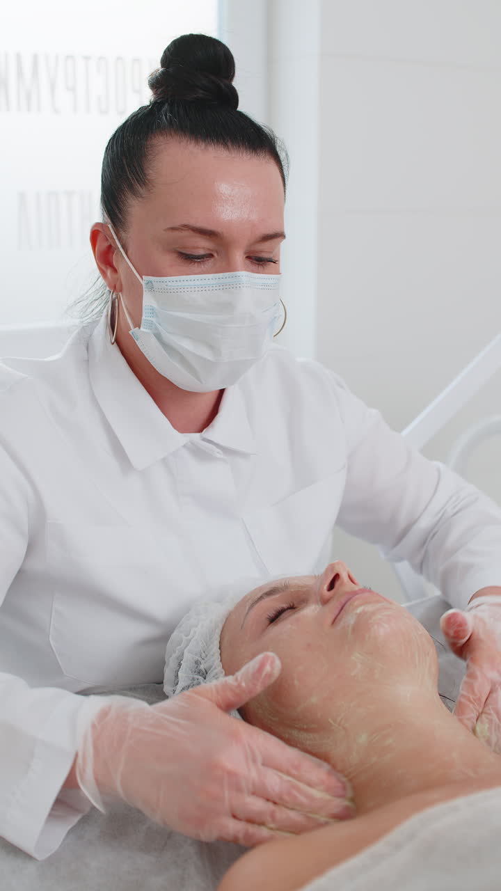 Cosmetologist applying moisturizing medical chemical peeling cream mask on young adult woman face