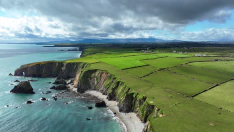 Drone-Estableciendo-Una-Toma-De-Una-Impresionante-Costa-Con-Campos-Verde-Esmeralda-Y-Montañas-En-El-Fondo,-El-Espectacular-Paisaje-De-Waterford,-Irlanda,-En-Primavera