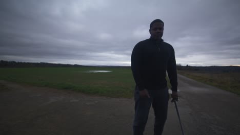 Modern-Black-Swordsman-Warrior-Awaiting-Battle-Opponent-in-Open-Field-Under-Grey-Sky,-Europe,-4K-|-Muscular,-Intimidating,-Shadow,-Figure,-Blurry,-Dark-Clothes,-Powerful,-Smile,-Lifting-Blade