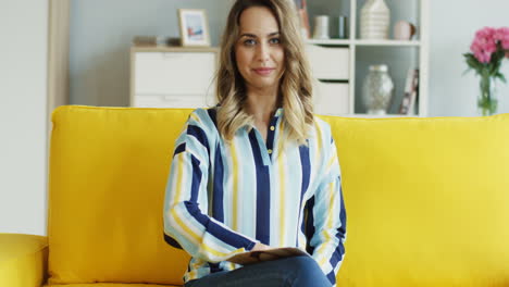 cheerful female blogger talking and showing thumbs up at camera while recording a video for her followers sitting on a yellow sofa at home