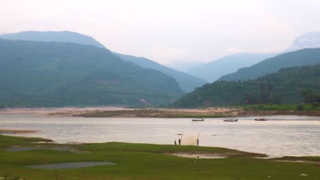 Wooden-boats-glide-on-a-river-in-Bholaganj,-Sylhet,-Bangladesh-India-border,-as-fishermen-work-beneath-clouds-on-a-hill