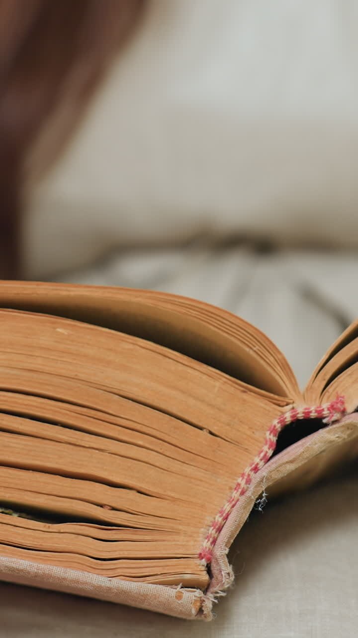 primo piano di un libro vintage sulle gambe di una donna caucasica che sfoglia delicatamente le pagine ingiallite con una rilegatura in pelle consumata, illuminazione calda e soffusa, texture tattile della carta, atmosfera intima e silenziosa, momento di studio tranquillo a casa
