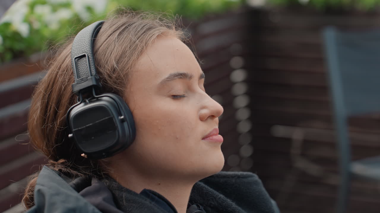 Giovane donna che si rilassa con le cuffie sulla terrazza panoramica sotto una leggera pioggerellina, occhi chiusi, capelli intrecciati, cappotto pesante, fioriere in legno con vegetazione, suono ambientale delicato, espressione serena, cinematografico