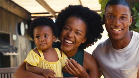 Portrait-Of-Parents-Playing-With-Young-Daughter-Sitting-On-Table-Outdoors-At-Home