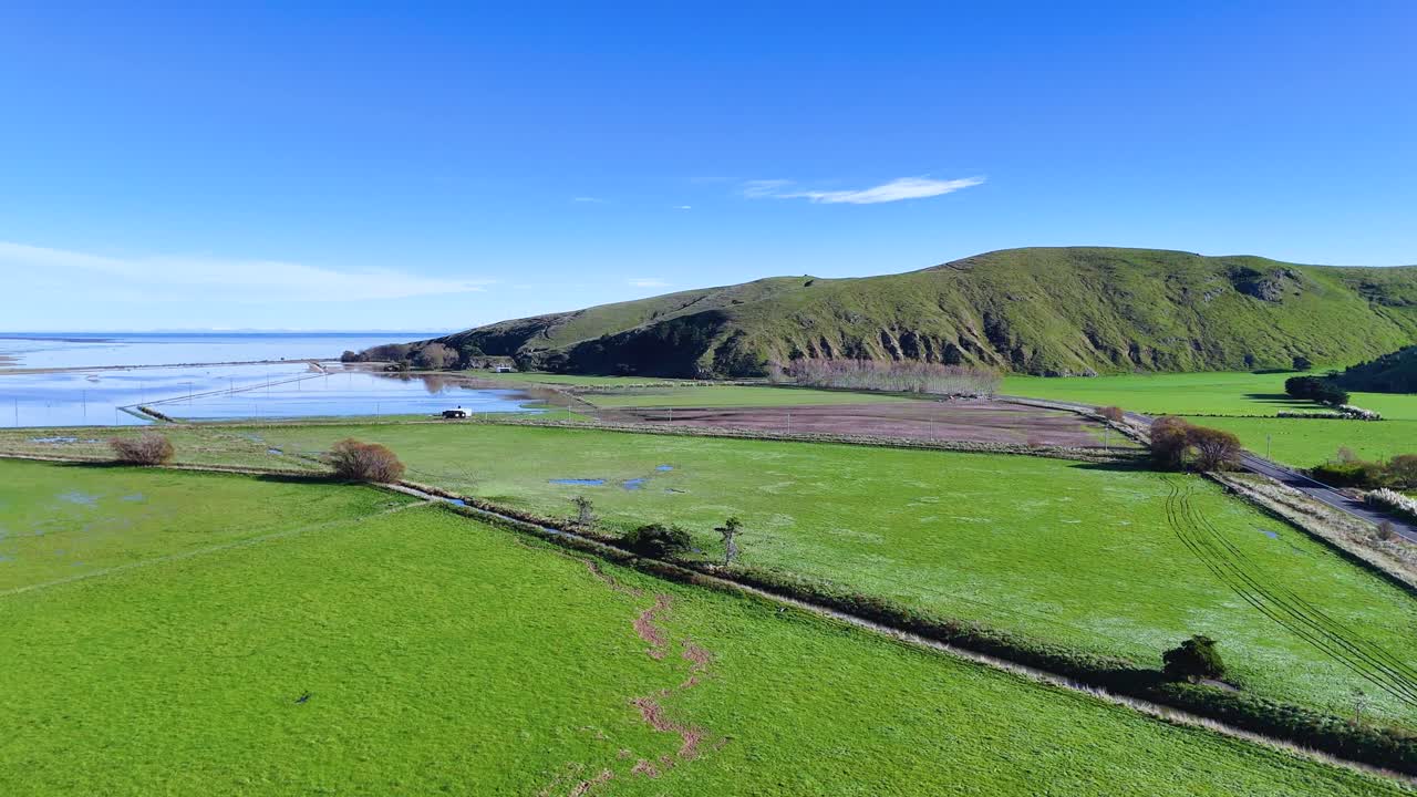 Aerial footage captures lush green fields and coastal hills under clear blue skies in Akaroa, New Zealand