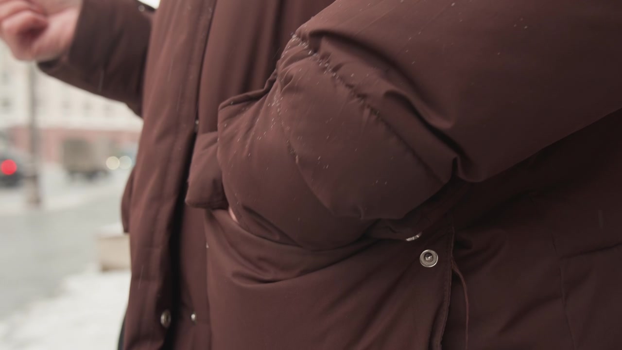 Torso focused motion scene shows person wearing brown padded jacket with right hand in pocket and left hand lifting forward, suggesting reaching or adjusting outerwear on city street