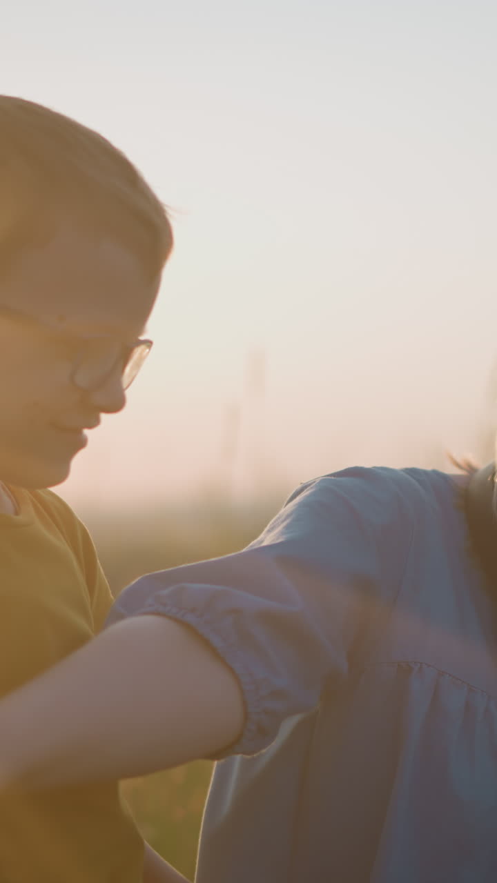 primer plano de una mujer con una túnica azul sentada en un campo cubierto de hierba al atardecer, tratando juguetón de detener a un niño vestido de amarillo de distraerla mientras un niño pequeño de blanco se sienta a su lado