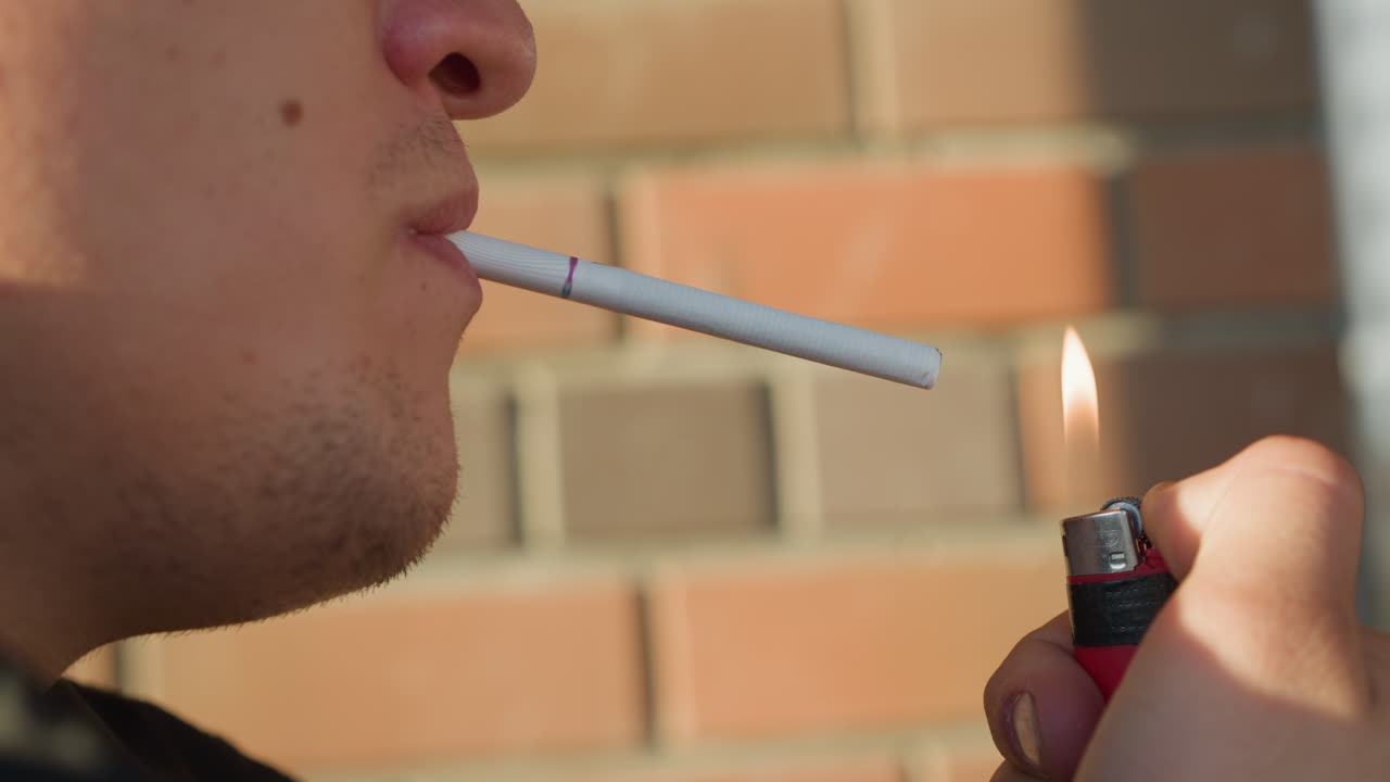 close up of boy holding cigarette in mouth with lit lighter positioned near cigarette tip, pausing in hesitation while standing against outdoor brick wall background during daylight
