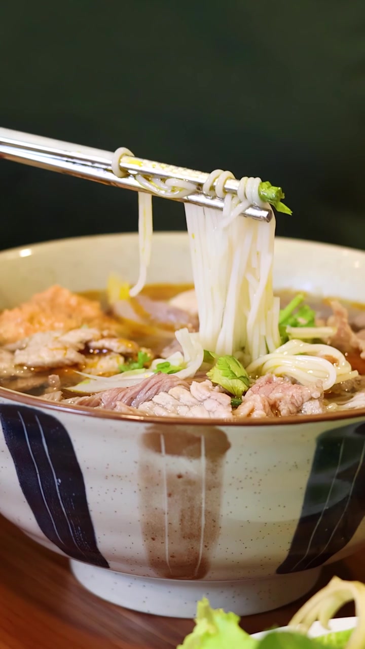 Một người dùng đũa kim loại gắp sợi phở từ một tô phở bò.