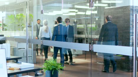 suited asian businessman rides skateboard through the corporate building hallway. stylish glass and concrete building with multicultural crowd of business people.