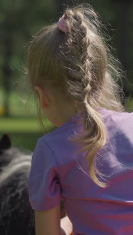 brave little girl with plait rides obedient pony with instructor along green lawn in resort park on sunny day backside view slow motion