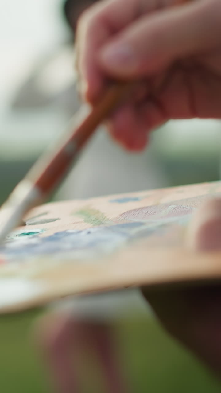 화가의 손이 팔레트를 들고 으로 페인트를 적용하는 클로즈업, 배경에 여성의 흐릿한 형상이 장면에 깊이를 더합니다.