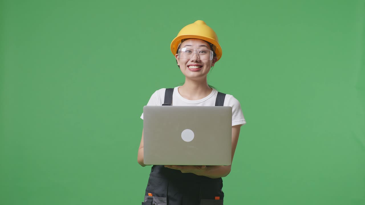 녹색 스크린 배경 스튜디오에 서 있는 아시아 여성 노동자가 노트북을 사용하고 을 가리키며 보호 안경과 안전 헬을 착용하고 있습니다.