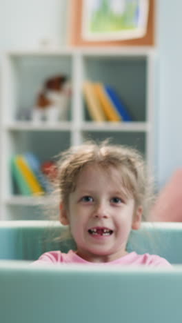 toothless preschooler throws plastic balls up. smiling girl has fun lying in dry pool at home. positive child experiences change of milk teeth to permanent