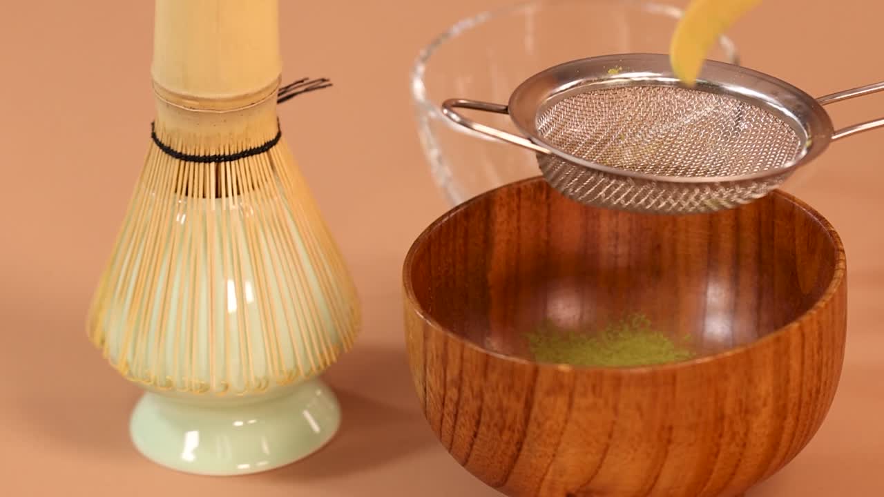 Lähikuva bambukauhasta siivilöimässä matcha-jauhetta metallisiivilän läpi puiseen kulhoon.