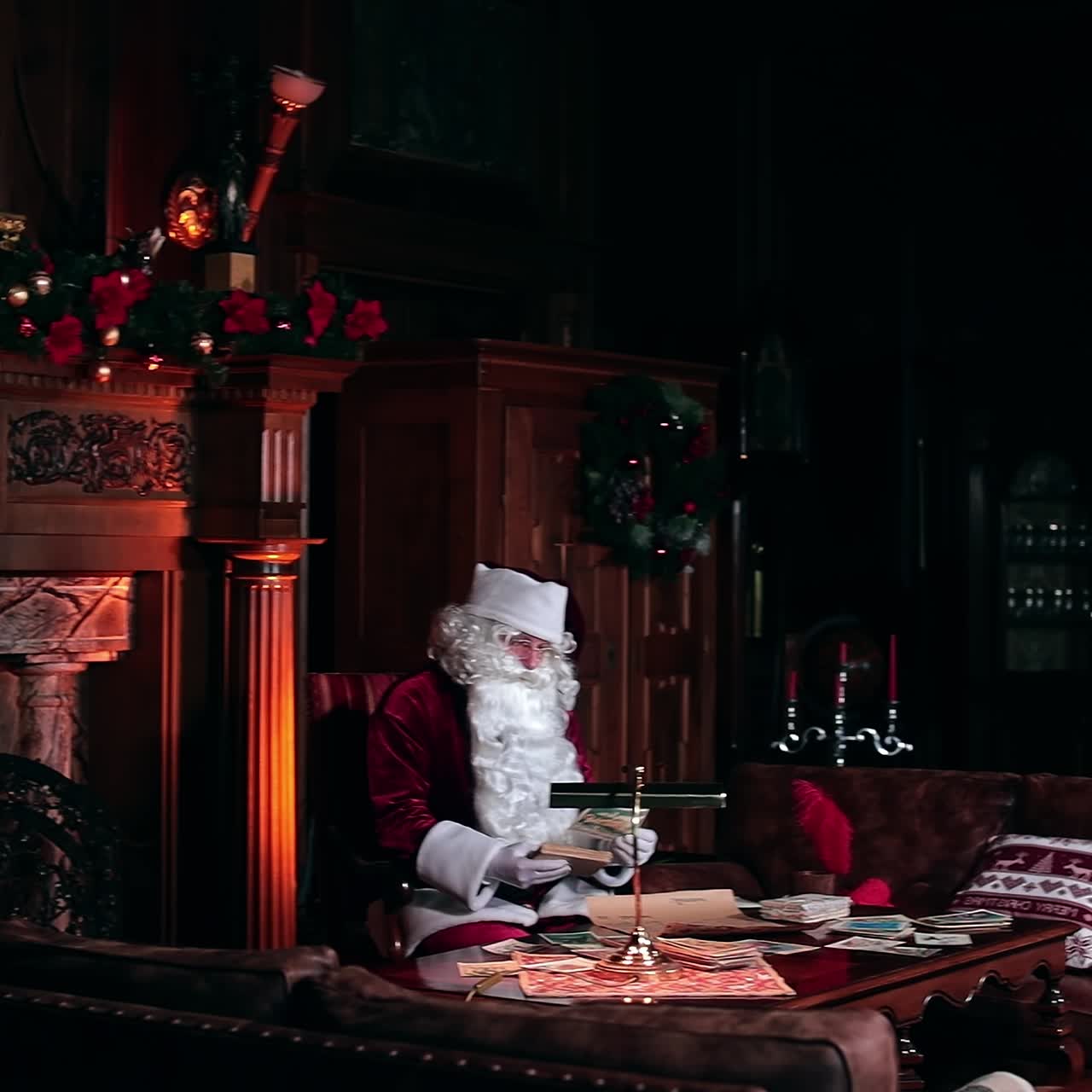 Beautiful cozy room with fireplace and old furniture. Santa Claus working sitting at the table looking through the cards