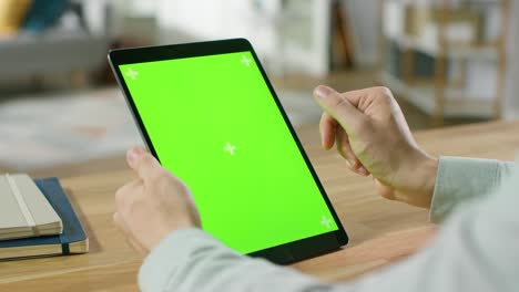 hombre usando gestos de la mano en pantalla de maqueta verde computadora tableta digital en modo retrato mientras está sentado en su escritorio. en el fondo acogedor salón.