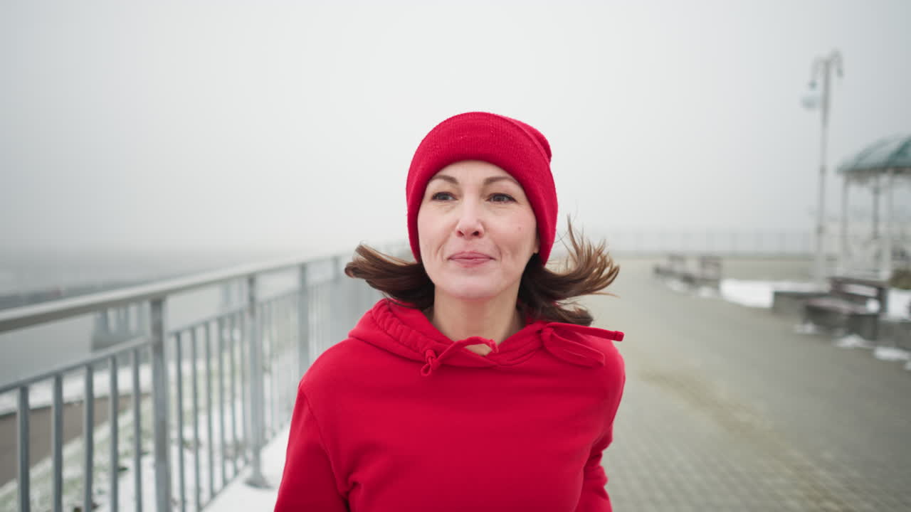primer plano de una mujer corriendo con gorra roja y chaqueta a lo largo de un camino nevado cerca de una barandilla de hierro, un parque de invierno sereno con bancos cerca, atmósfera de niebla, puente lejano y poste de luz