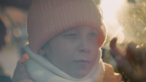 close-up of a young girl wearing a pink knit hat and white scarf, sitting inside a car, drawing on the window glass with her finger, one eye closed