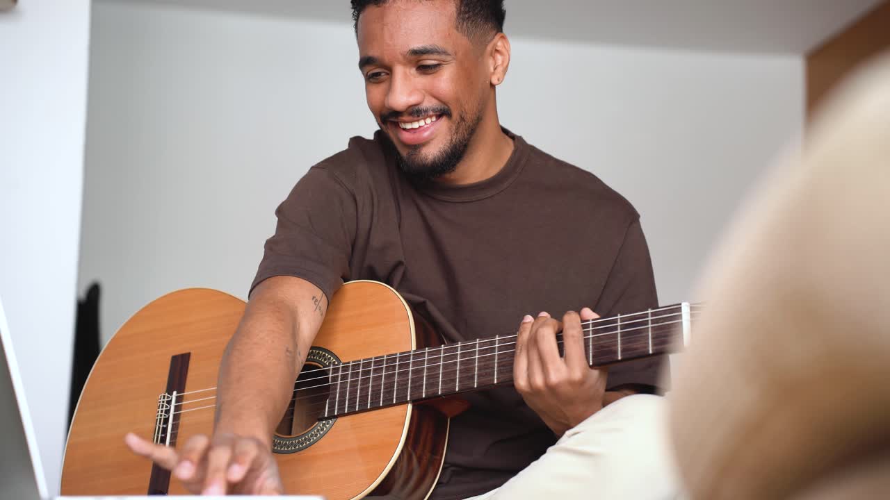 hombre étnico alegre tocando la guitarra con una computadora portátil en el sofá