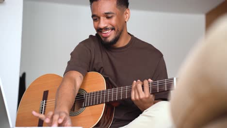 hombre étnico alegre tocando la guitarra con una computadora portátil en el sofá