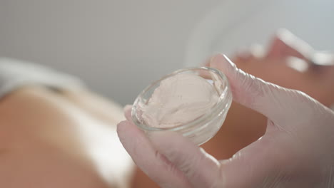 gloved body work expert hand holding transparent silicone cup loaded with rich white facial cream, soft focus spa environment, crisp closeup showcasing treatment product in serene clinical wellness