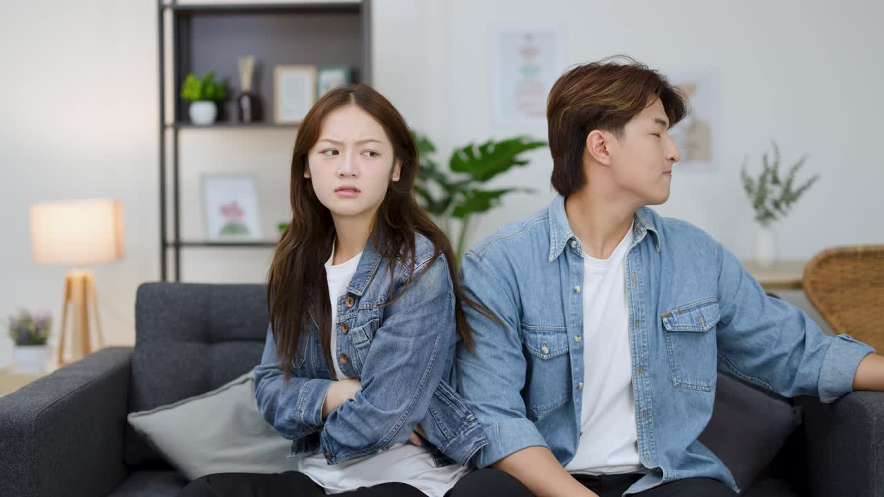 คู่รักชาวเอเชียหนุ่มสาวกำลังโต้เถียงกันบนโซฟา จนในที่สุดก็หันหน้าหนีกันด้วยความเงียบงันอย่างหงุดหงิด
