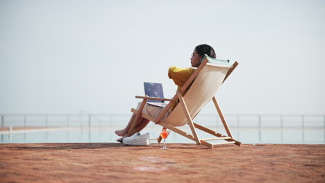 woman relaxing in a beach chair with laptop and cocktail by the pool