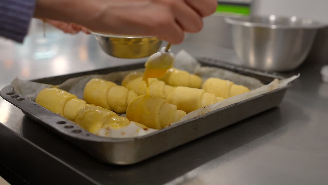 Mani che spennellano dei croissant crudi con doratura all'uovo in cucina.