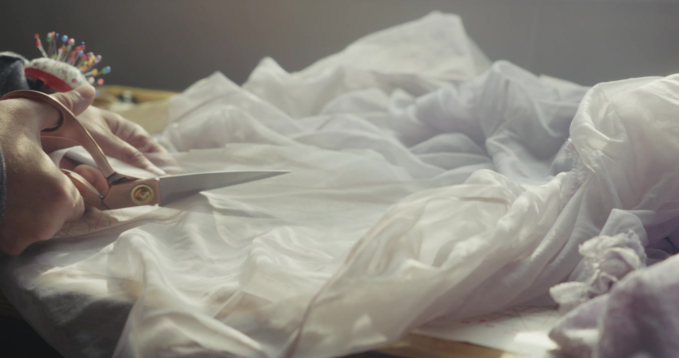 Primo piano di uno stilista di moda che taglia un tessuto di tulle bianco semitrasparente su un tavolo da lavoro in studio, evidenziando la sartoria di precisione e l'artigianato tessile.
