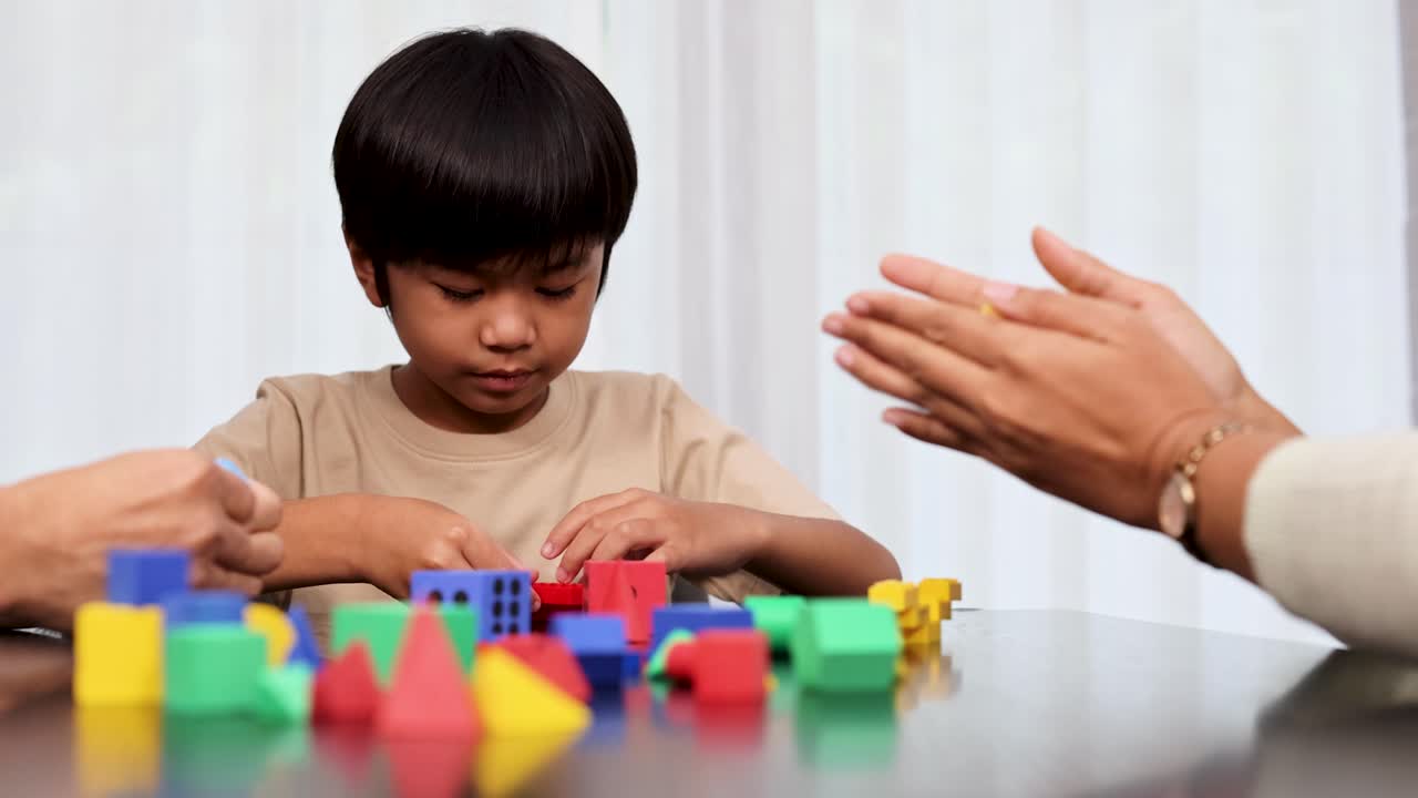 Un giovane ragazzo asiatico e i suoi genitori interagiscono con blocchi giocattolo colorati e dadi su un tavolo in un ambiente interno luminoso e delicatamente illuminato. La telecamera rimane ferma, catturando momenti giocosi ed educativi.