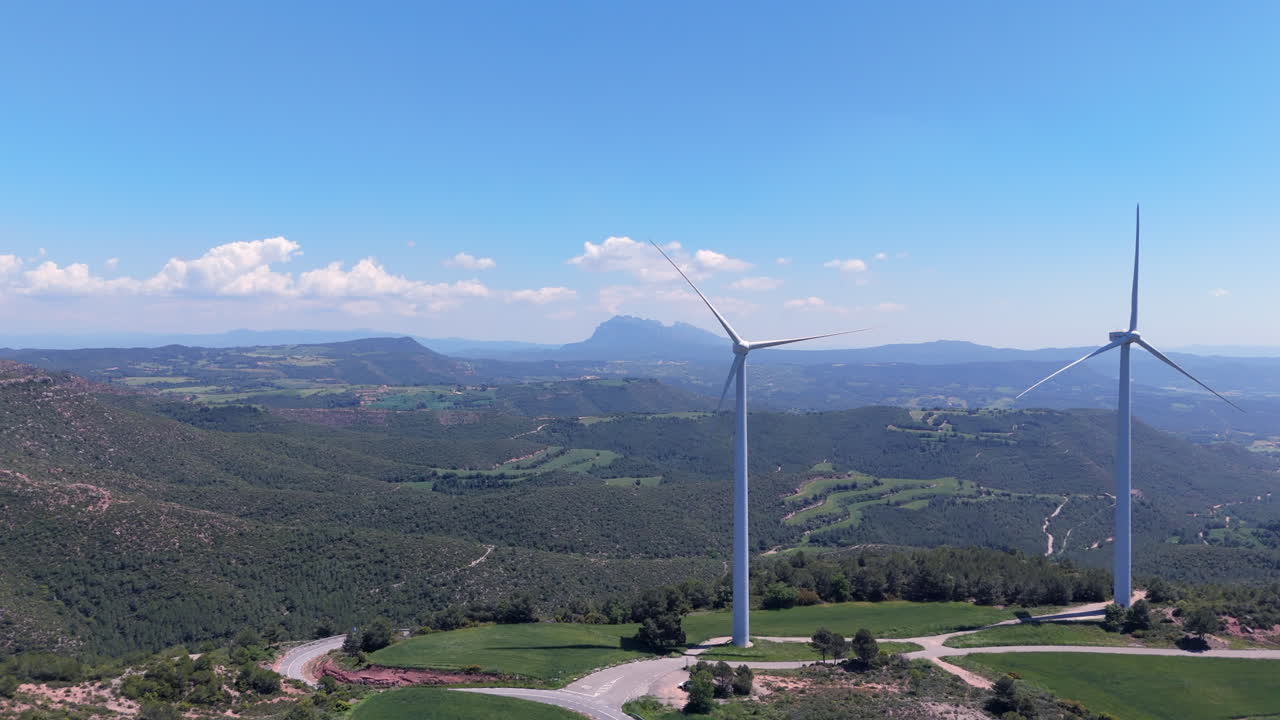 Lateral drone traveling over a mountain landscape with two large wind turbines under a bright blue sky. Renewable energy infrastructure surrounded by nature and distant ridges