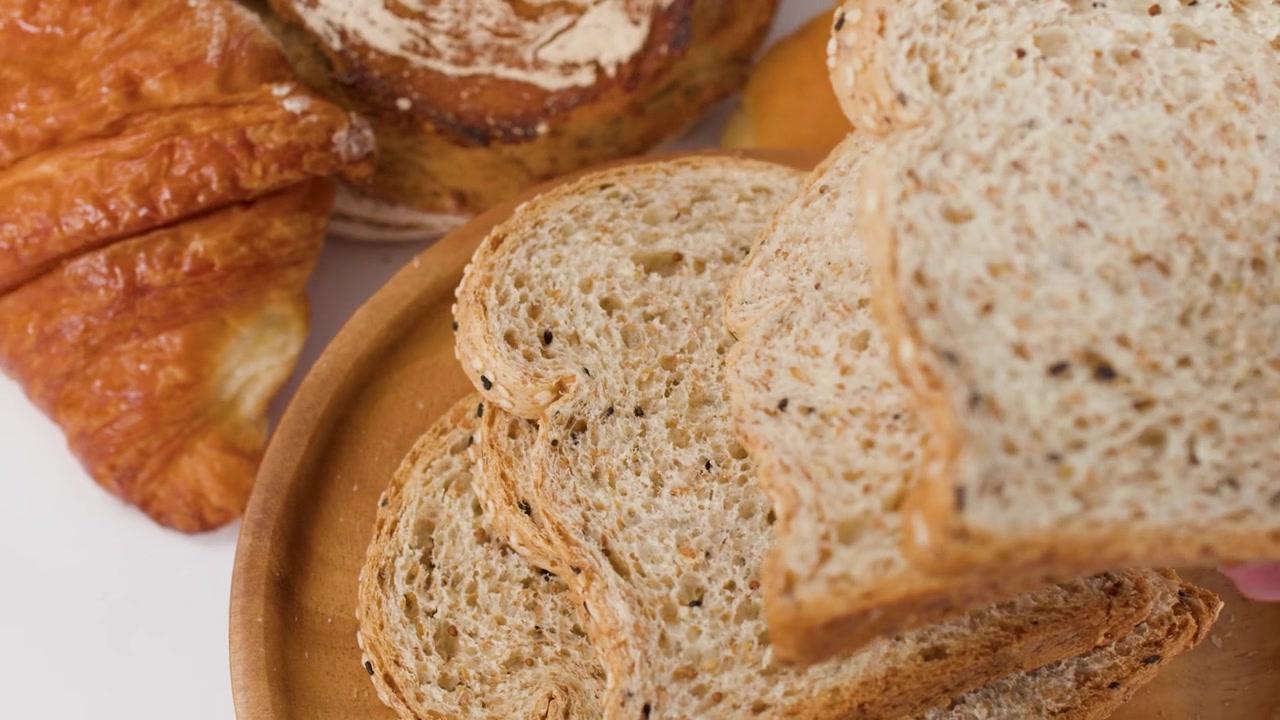 Close-up of hands arranging sliced whole wheat bread among assorted bakery items on white background.