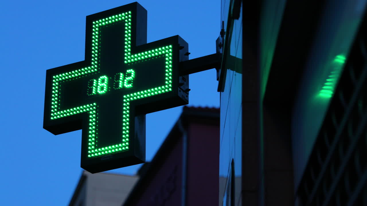 Zoom su farmacie e simbolo internazionale illuminato appeso all'angolo di un edificio con un orologio scintillante al centro di una croce verde che mostra una farmacia, ripreso in 4K a 60fps
