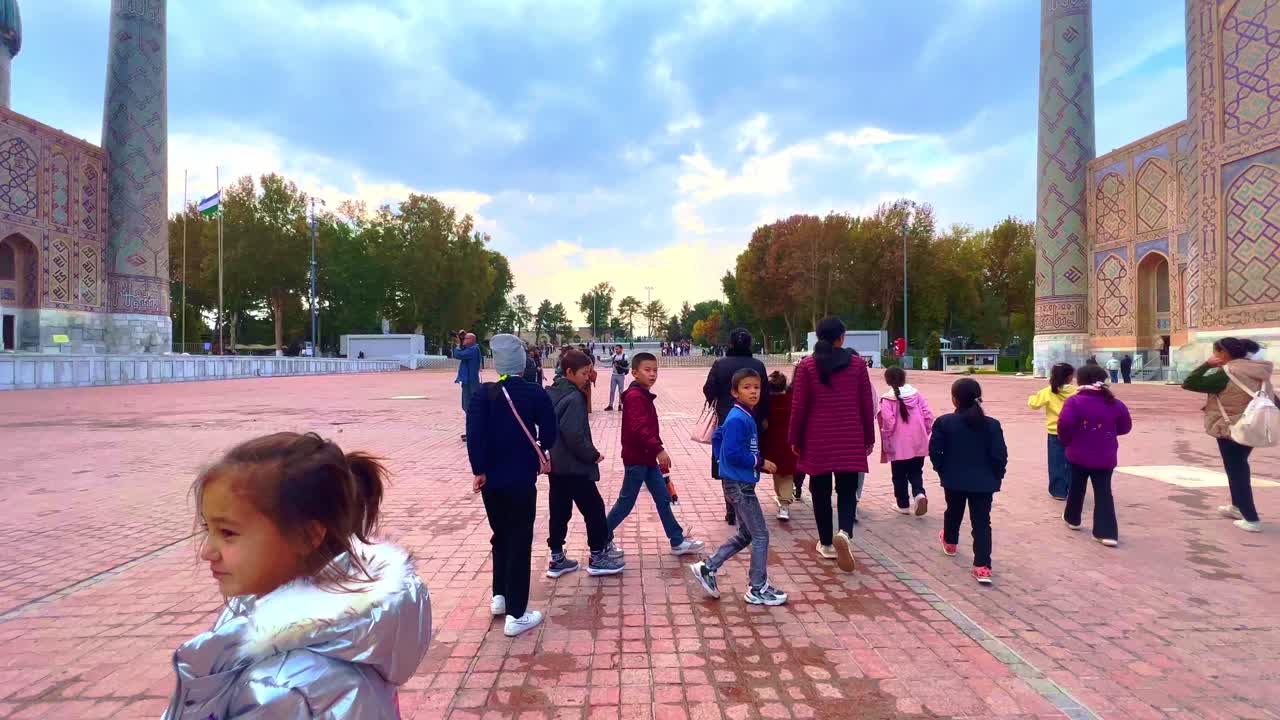 People and tourists exploring the vast historical Registan Square in Samarkand, featuring grand Islamic architecture under a cloudy sky.