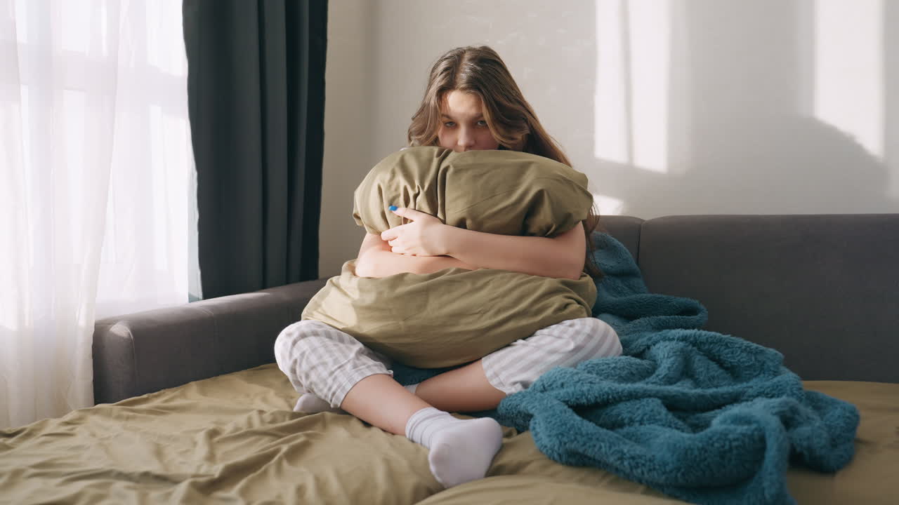 White Teenage Student Clutching Pillow On Sofa, Crosslegged With Striped Pajamas And Knitted Blanket, Sunlight Falling Through Window, Thoughtful Expression And Quiet Reflection In Domestic Interior