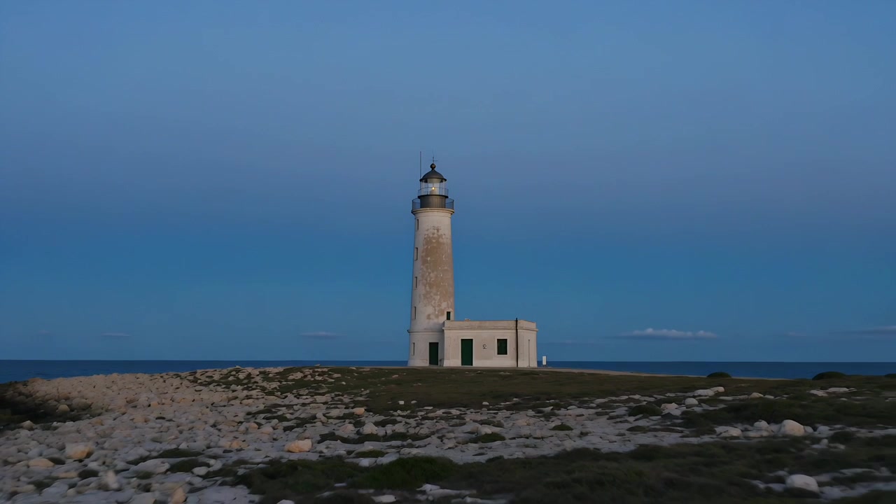 La telecamera si allontana, rivelando la torre del faro e l'edificio del guardiano su un promontorio roccioso al crepuscolo.