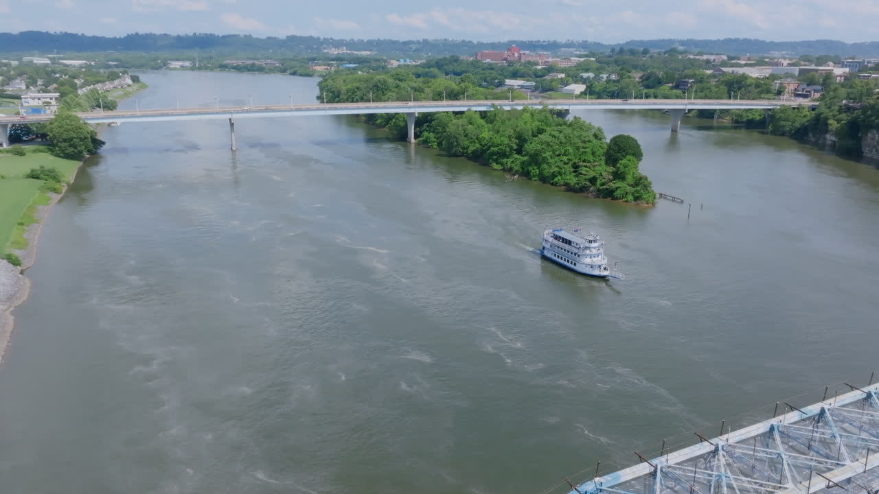 An aerial view of the Tennessee River in Chattanooga, TN, shows a riverboat cruising near a bridge and a lush island under a partly cloudy sky