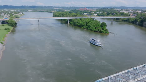 an aerial view of the tennessee river in chattanooga, tn, shows a riverboat cruising near a bridge and a lush island under a partly cloudy sky