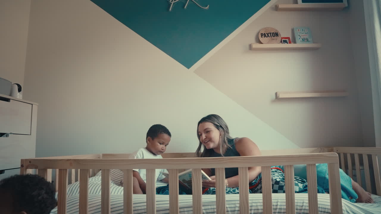 Hispanic mom reads bedtime story to toddler in bed as Black dad tidies the bedroom. Infant sits in foreground, capturing teamwork, siblings, and authentic family life