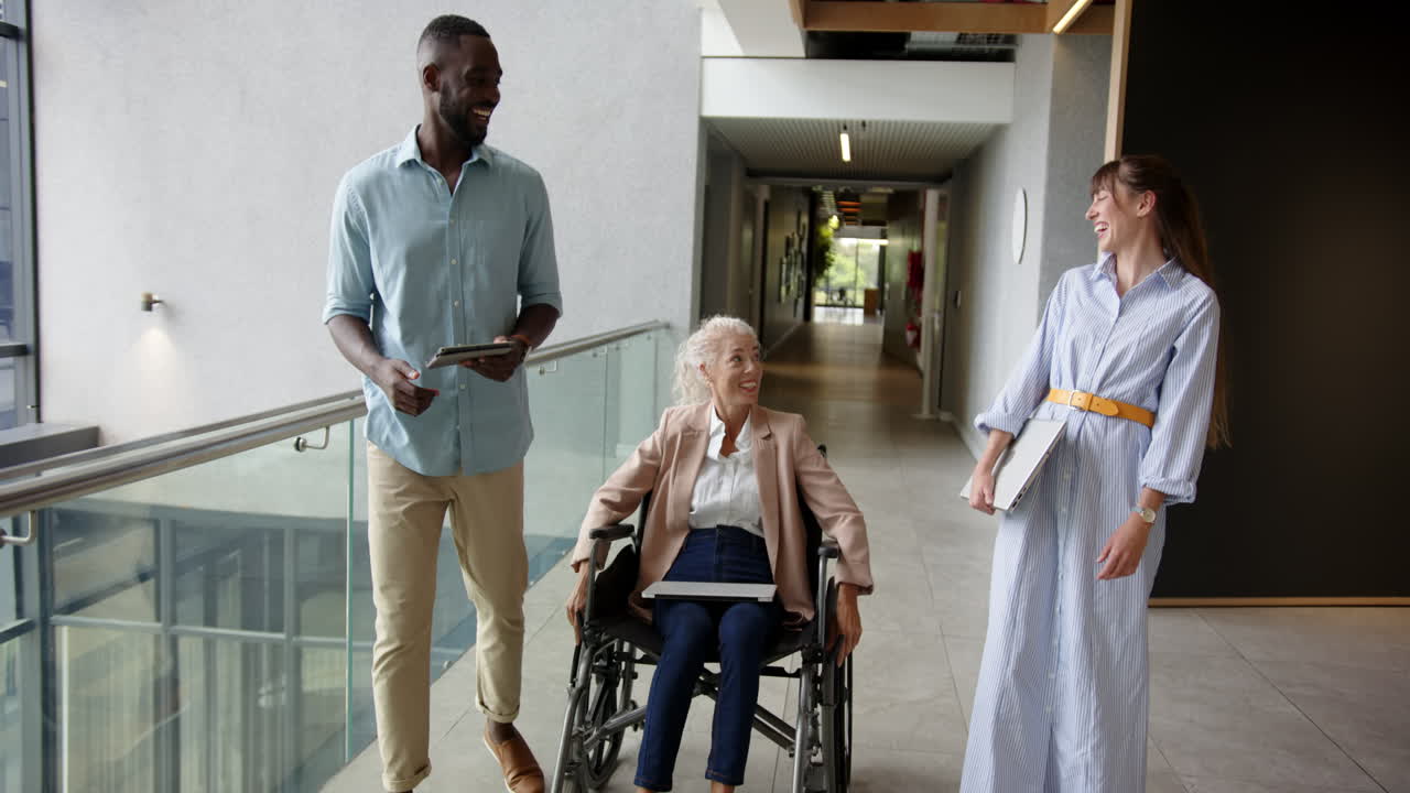 Walking in office, diverse colleagues one using wheelchair discussing work with woman in wheelchair