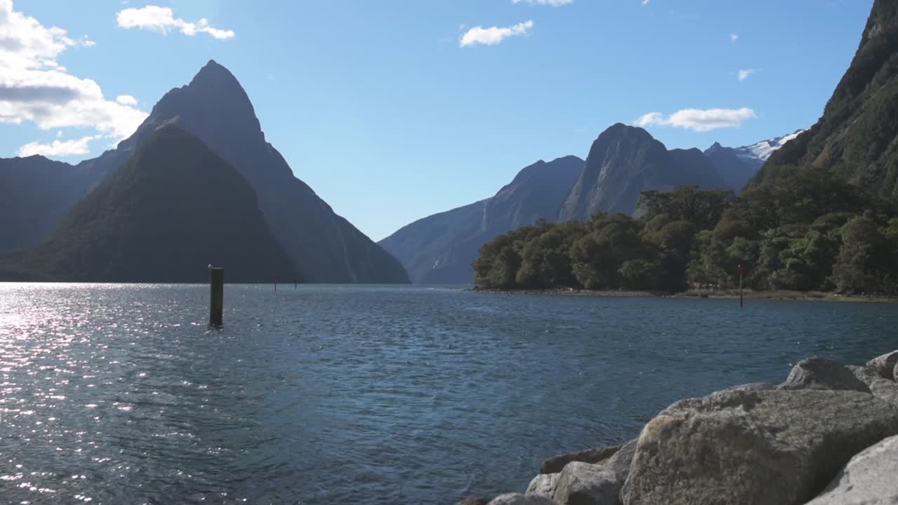 Vista diurna del Milford Sound in una giornata di cielo sereno. L'acqua in primo piano, il Mitre Peak e altre montagne in lontananza.