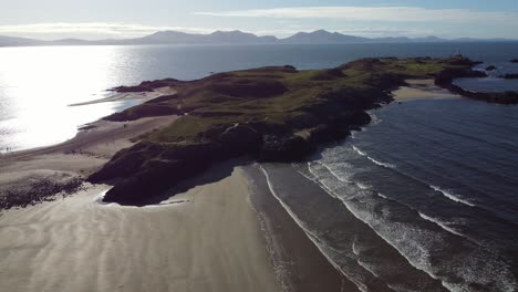 wide aerial view circling ynys llanddwyn welsh island with shimmering ocean and misty snowdonia mountain range across the sunrise skyline