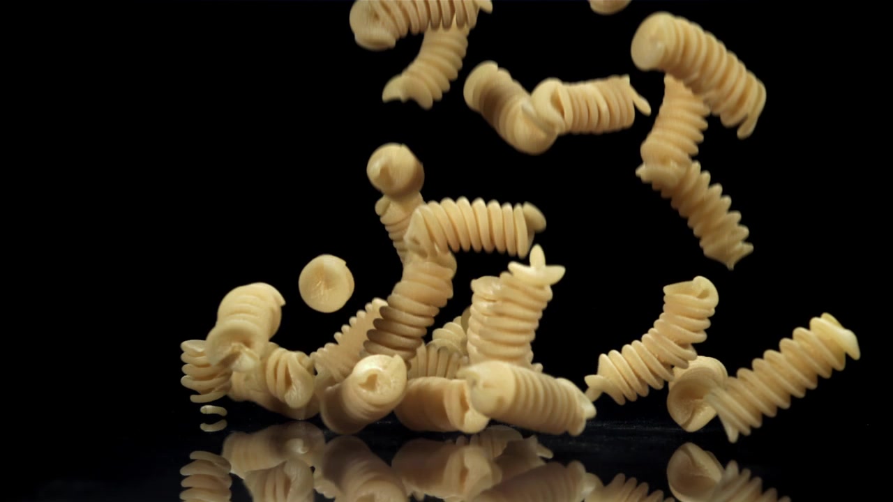 Pasta fusilli secca che cade su una superficie nera riflettente.