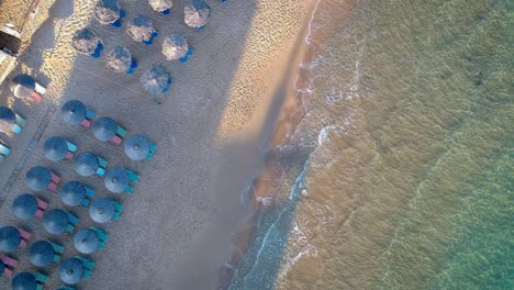 Vista-Aérea-De-Arriba-Hacia-Abajo-Sobre-Las-Olas-Rompiendo-En-La-Playa-Con-Tumbonas-Vacías-Con-Sombrillas-Azules