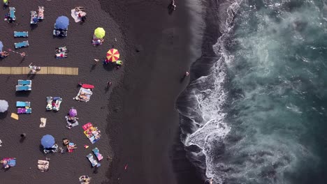 Touristen,-Die-Während-Der-Ferienzeit-In-Der-Luftaufnahme-Von-Oben-Nach-Unten-Am-überfüllten-Tropischen-Strand-Mit-Schwarzem-Sand-Und-Sonnenschirm-Spazieren-Gehen