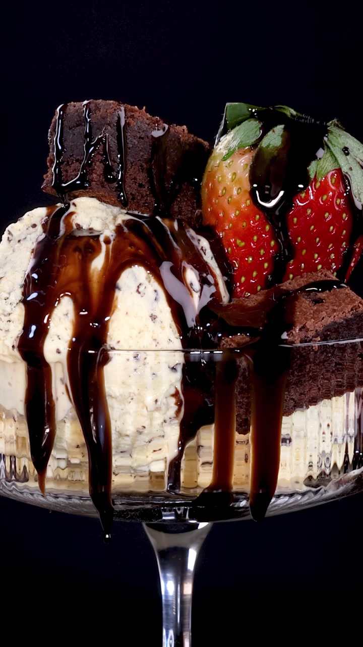 Salsa al cioccolato a rallentatore che si versa su gelato alla vaniglia, brownies e una fragola fresca in una coppetta di vetro per dessert, su uno sfondo scuro con illuminazione drammatica.