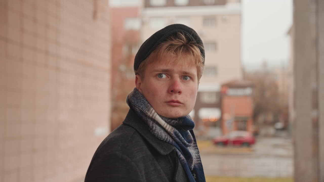 Young creator on city balcony, contemplative scout filmmaker scouting location, scarf and beanie, coat textures, soft overcast light, shallow depth, distant traffic and storefront blur, thoughtful